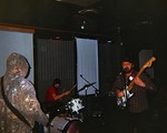 A grainy candid photo shows three-piece band Invisible Hand performing in a dark room with curtains drawn over the windows. 
