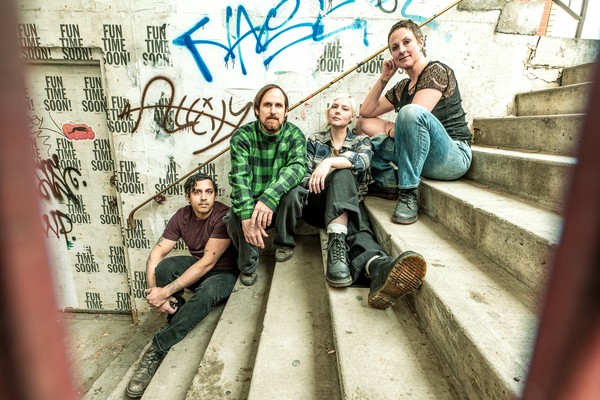 A colour photo shows four-piece group innerouter sitting outside on concrete stairs, with each member on a different step.The setting that resembles the enrtrance to an old train station, graffiti fills the concrete wall behind them.
