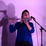 A colour photo shows artist JJL playing flute in front of a lavender wall. They are wearing a blue shirt and have their eyes closed, playing passionately. 