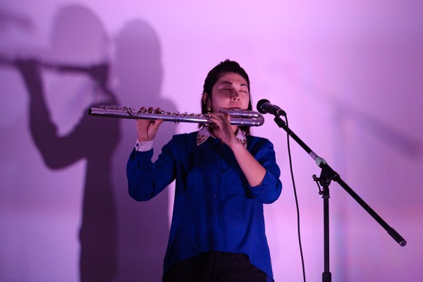 A colour photo shows artist JJL playing flute in front of a lavender wall. They are wearing a blue shirt and have their eyes closed, playing passionately. 