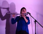 A colour photo shows artist JJL playing flute in front of a lavender wall. They are wearing a blue shirt and have their eyes closed, playing passionately. 