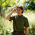 A colour photo shows artist Jo Passed standing in a green field with long grass and leady trees. They are wearing as dark green t-shirt and seen tucking their brown hair behind their ear. 