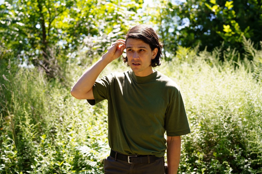 A colour photo shows artist Jo Passed standing in a green field with long grass and leady trees. They are wearing as dark green t-shirt and seen tucking their brown hair behind their ear. 
