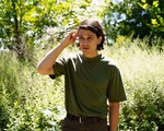 A colour photo shows artist Jo Passed standing in a green field with long grass and leady trees. They are wearing as dark green t-shirt and seen tucking their brown hair behind their ear. 