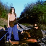 Duo Kali Horse are seen outside on a railroad track with green leafy tress on either side and a blue sky above. One member stand withs a guitar strapped on, and the other is seen sitting on tracks with their legs outstrecthed. 