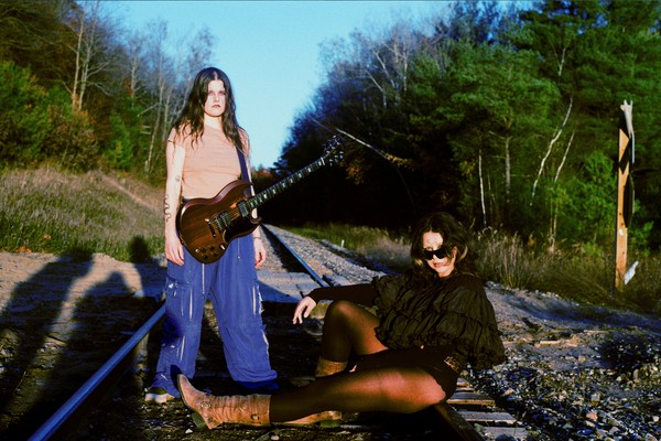 Duo Kali Horse are seen outside on a railroad track with green leafy tress on either side and a blue sky above. One member stand withs a guitar strapped on, and the other is seen sitting on tracks with their legs outstrecthed. 