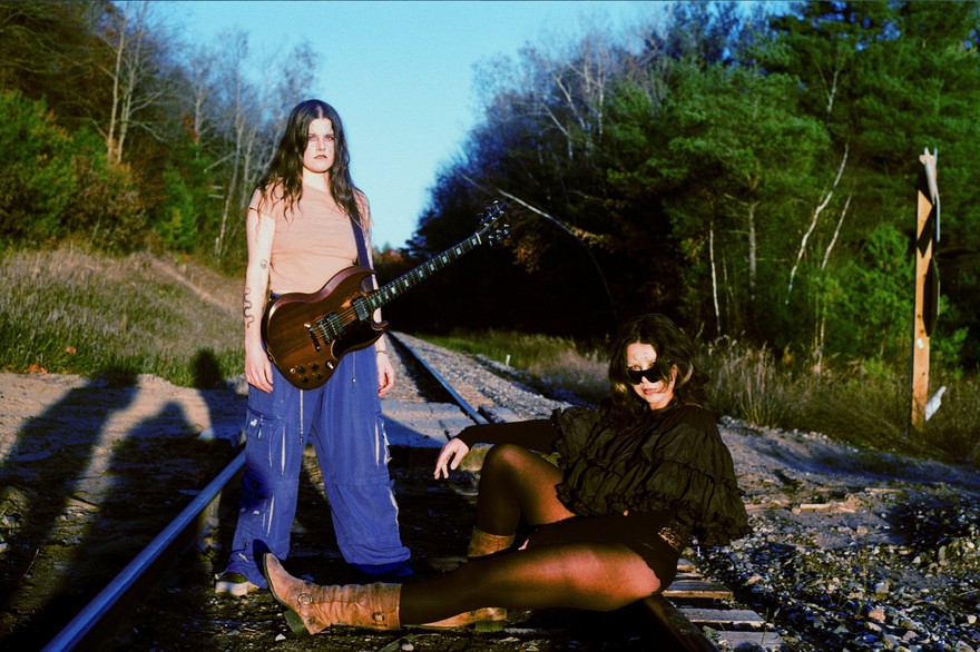 Duo Kali Horse are seen outside on a railroad track with green leafy tress on either side and a blue sky above. One member stand withs a guitar strapped on, and the other is seen sitting on tracks with their legs outstrecthed. 