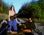 Duo Kali Horse are seen outside on a railroad track with green leafy tress on either side and a blue sky above. One member stand withs a guitar strapped on, and the other is seen sitting on tracks with their legs outstrecthed. 