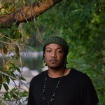 Artist K-Riz is shown standing outside with green leafy trees around them and a body of water in the distance. They are wearing a black t-shirt and a dark green toque. 