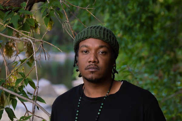 Artist K-Riz is shown standing outside with green leafy trees around them and a body of water in the distance. They are wearing a black t-shirt and a dark green toque. 