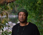 Artist K-Riz is shown standing outside with green leafy trees around them and a body of water in the distance. They are wearing a black t-shirt and a dark green toque. 