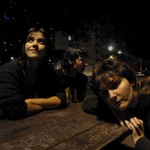 Three members of the group Kingdom of Birds are shown sitting at a brown picnic table at nighttime with various buildings shown behind them. 