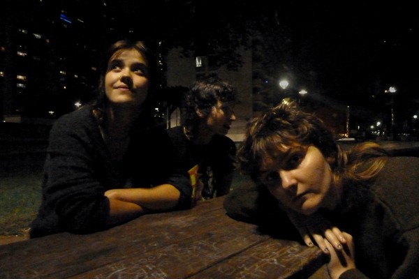 Three members of the group Kingdom of Birds are shown sitting at a brown picnic table at nighttime with various buildings shown behind them. 