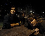 Three members of the group Kingdom of Birds are shown sitting at a brown picnic table at nighttime with various buildings shown behind them. 