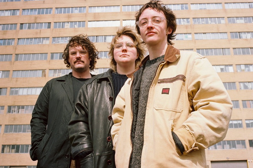 A colour photos shows three-piece group knitting standing outside in front of a large brick building with many windows.