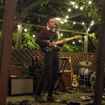 Artist l.n. baba is shown performing on an outdoor stage with tress behind and above, and string lights hanging from the wooden structure above. They are playing guitar and singing, while various amps, guitars and a drumkit is shown around them. 
