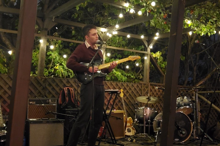Artist l.n. baba is shown performing on an outdoor stage with tress behind and above, and string lights hanging from the wooden structure above. They are playing guitar and singing, while various amps, guitars and a drumkit is shown around them. 