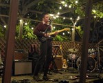 Artist l.n. baba is shown performing on an outdoor stage with tress behind and above, and string lights hanging from the wooden structure above. They are playing guitar and singing, while various amps, guitars and a drumkit is shown around them. 