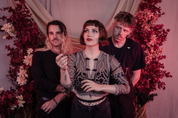 A colour photo shows three members of Laura Hickli standing in front of a pink curtain with red and pink plants and flowers shown on both sides of them. 