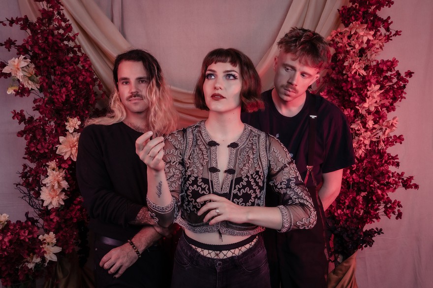 A colour photo shows three members of Laura Hickli standing in front of a pink curtain with red and pink plants and flowers shown on both sides of them. 