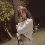 Singer-songwriter Lennie Rayen is shown outside leaning against a brown wodden pole at nighttime. They are wearing a white blouse and white pants. Trees and a green field are seen behind them. 