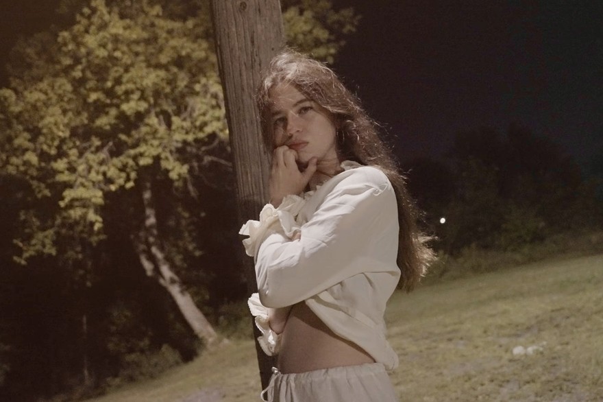 Singer-songwriter Lennie Rayen is shown outside leaning against a brown wodden pole at nighttime. They are wearing a white blouse and white pants. Trees and a green field are seen behind them. 