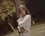 Singer-songwriter Lennie Rayen is shown outside leaning against a brown wodden pole at nighttime. They are wearing a white blouse and white pants. Trees and a green field are seen behind them. 