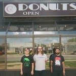 A grainy colour photo shows three-piece LOBOTOMITE standing outside of a building with mirrored glass behind them showing the reflection of the photgrapher and the street behind them. An old sign above reads "Donuts. Open."
