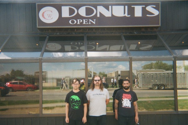 A grainy colour photo shows three-piece LOBOTOMITE standing outside of a building with mirrored glass behind them showing the reflection of the photgrapher and the street behind them. An old sign above reads "Donuts. Open."