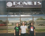 A grainy colour photo shows three-piece LOBOTOMITE standing outside of a building with mirrored glass behind them showing the reflection of the photgrapher and the street behind them. An old sign above reads "Donuts. Open."