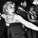 A black and white photo shows performer Lulu Lamontagne in a black corset holding a microphone and stretching the cord in front of them with their other hand. Two other members of the band are seen performing behind them. 