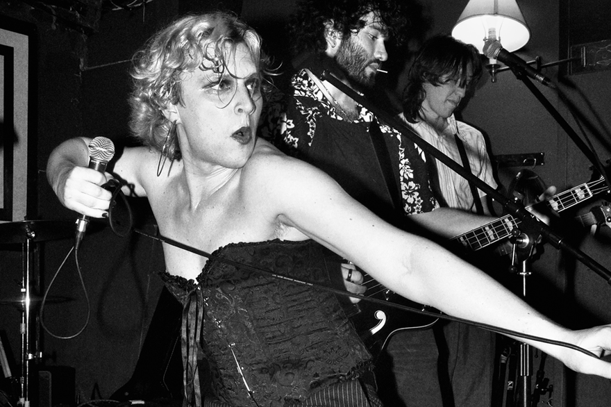 A black and white photo shows performer Lulu Lamontagne in a black corset holding a microphone and stretching the cord in front of them with their other hand. Two other members of the band are seen performing behind them. 