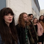 A colour photo of five-piece band Lushings shows them standing outside of a building leaning up againt the wall. The front person is shown in focus at the front with all the members standing behind them gradually getting less out of focus. 