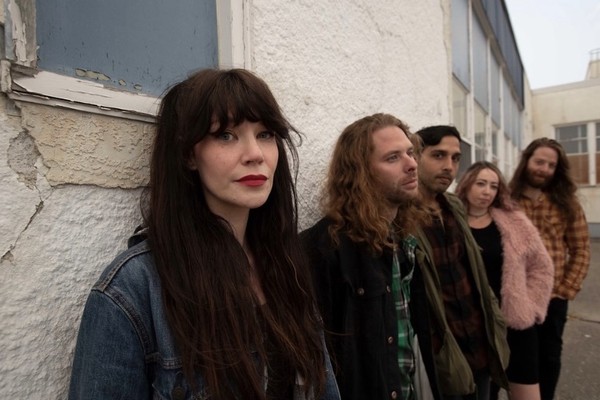A colour photo of five-piece band Lushings shows them standing outside of a building leaning up againt the wall. The front person is shown in focus at the front with all the members standing behind them gradually getting less out of focus. 