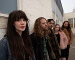 A colour photo of five-piece band Lushings shows them standing outside of a building leaning up againt the wall. The front person is shown in focus at the front with all the members standing behind them gradually getting less out of focus. 