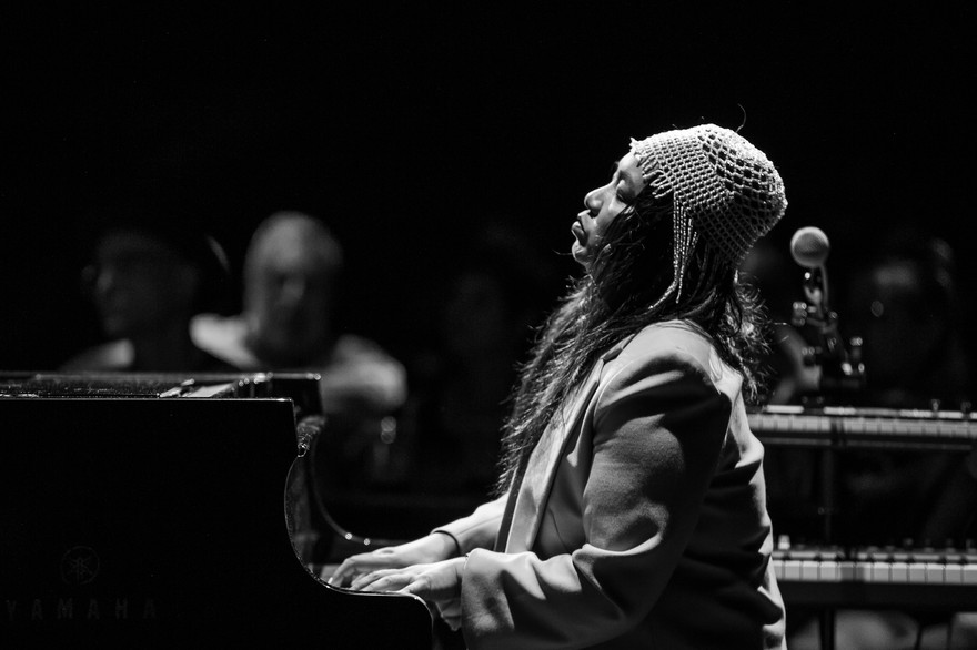 A black and white photo shows artist Mary Ancheta playing a black piano with an out of focus crowd shown behind them. 