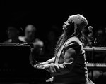 A black and white photo shows artist Mary Ancheta playing a black piano with an out of focus crowd shown behind them. 