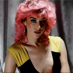 A close-up colour photo shows artist Maryze standing in front of a grey curtain. They have bright pink wavy hair, bright pink lipstick, and a black a yellow lowcut top. 