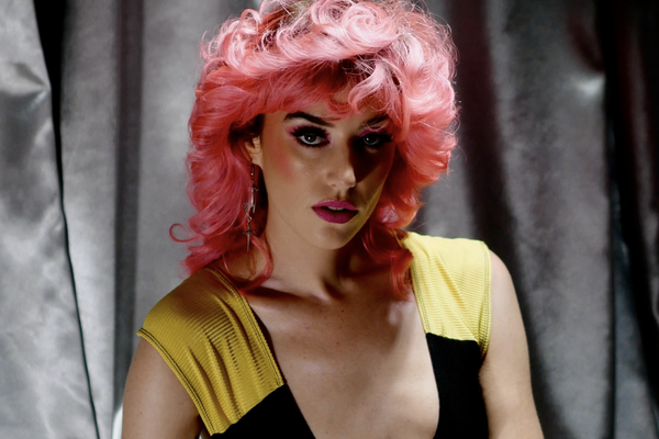 A close-up colour photo shows artist Maryze standing in front of a grey curtain. They have bright pink wavy hair, bright pink lipstick, and a black a yellow lowcut top. 
