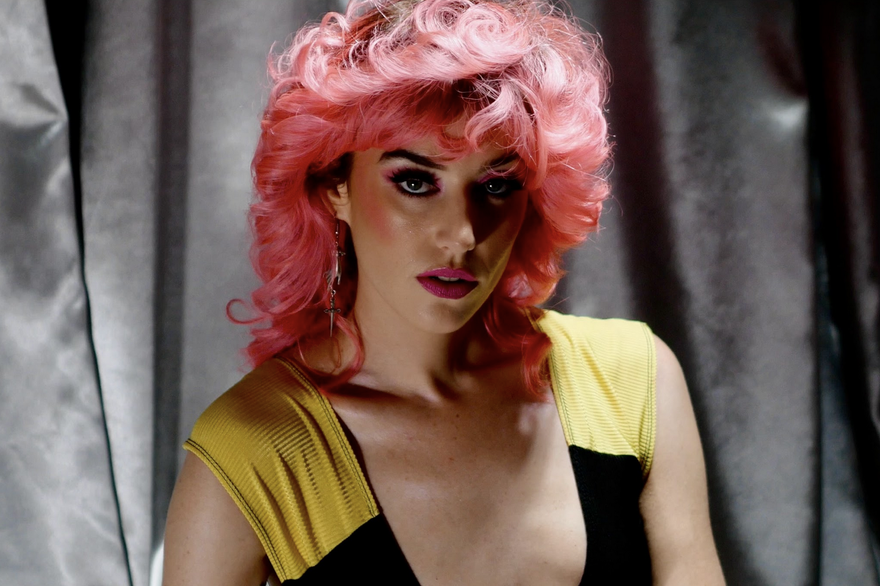A close-up colour photo shows artist Maryze standing in front of a grey curtain. They have bright pink wavy hair, bright pink lipstick, and a black a yellow lowcut top. 