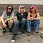 A colour photo shows young three-piece band Medusas sitting on a curb outside of a brick building looking tough at the camera. 