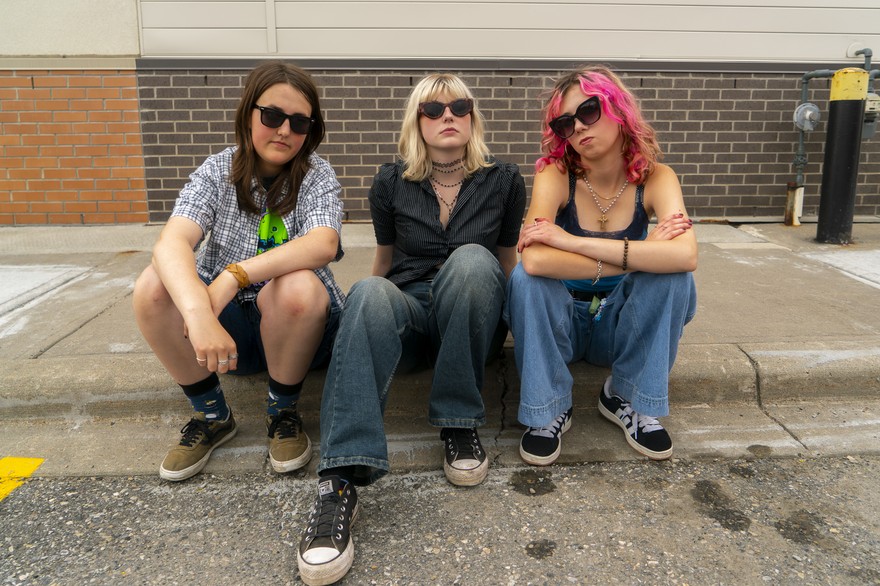 A colour photo shows young three-piece band Medusas sitting on a curb outside of a brick building looking tough at the camera. 