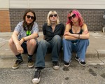 A colour photo shows young three-piece band Medusas sitting on a curb outside of a brick building looking tough at the camera. 