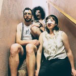 Three members of the band Minot are shown sitting on a narrow indoor staircase. One member has a bandage on their nose, another on their forehead, and another over their eye. 