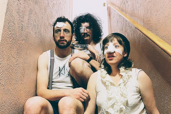 Three members of the band Minot are shown sitting on a narrow indoor staircase. One member has a bandage on their nose, another on their forehead, and another over their eye. 