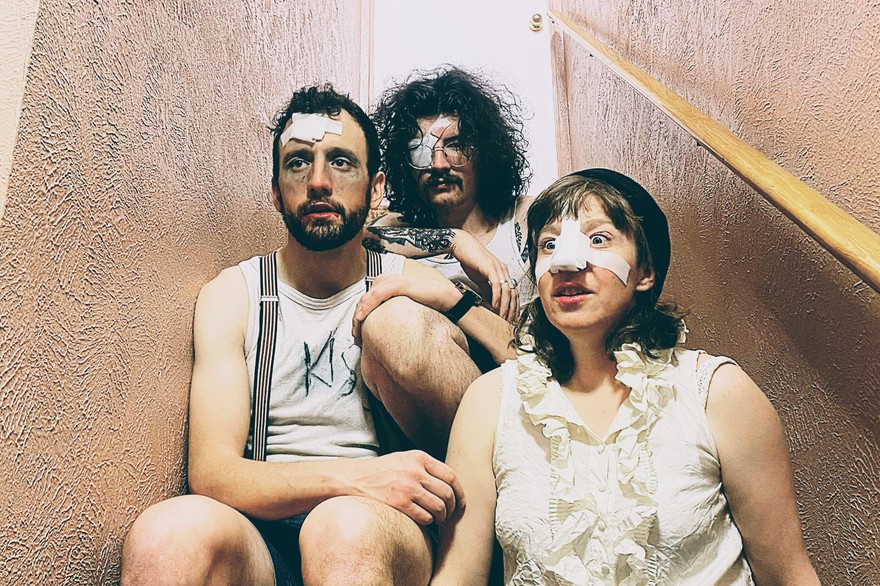 Three members of the band Minot are shown sitting on a narrow indoor staircase. One member has a bandage on their nose, another on their forehead, and another over their eye. 