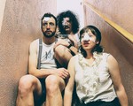 Three members of the band Minot are shown sitting on a narrow indoor staircase. One member has a bandage on their nose, another on their forehead, and another over their eye. 