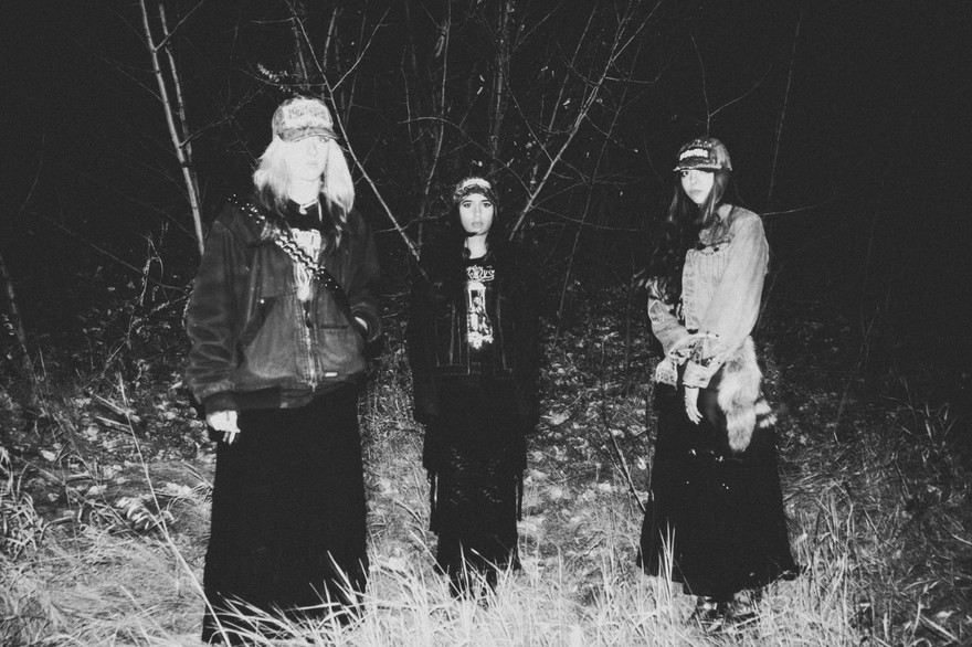 A fuzzy black and white photo shows three-piece band standing outside in a field at night with long grass and trees behind them. 