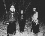 A fuzzy black and white photo shows three-piece band standing outside in a field at night with long grass and trees behind them. 