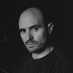 A close-up black and white photo shows artist Nick Schofield in a black t-shirt against a black background looking directly at the camera. 